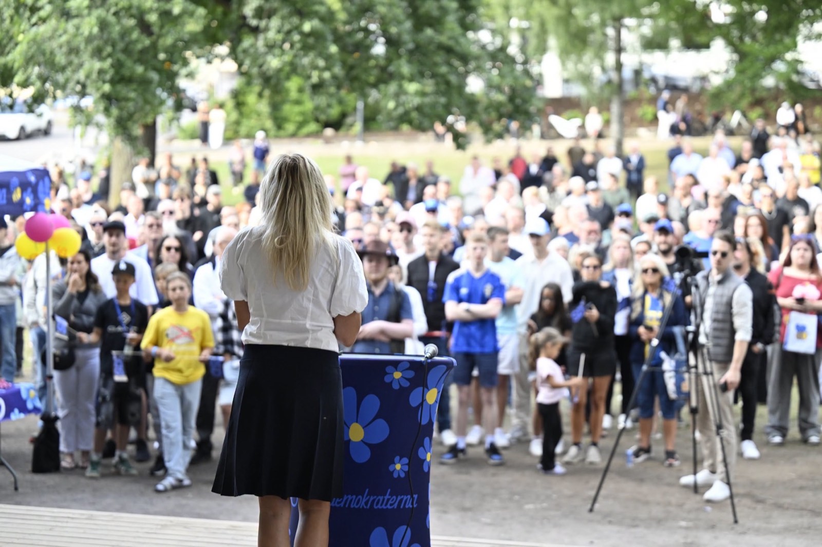 Linda Lindberg står på en scen med ryggen mot kameran och talar till en stor publik i en park.